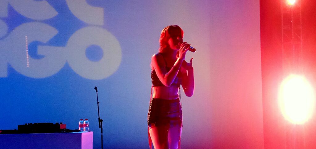 Female singer performing on a colorful stage, microphone in hand, with blue to pink lighting and DJ gear on a table nearby.