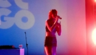 Female singer performing on a colorful stage, microphone in hand, with blue to pink lighting and DJ gear on a table nearby.