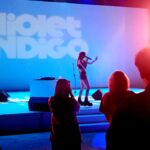 Singer on stage with a microphone under blue lighting; audience silhouettes in foreground and a backdrop reading 'Violet Indigo'.
