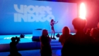 Singer on stage with a microphone under blue lighting; audience silhouettes in foreground and a backdrop reading 'Violet Indigo'.