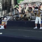 Mime with white makeup, striped shirt and yellow vest sits on stage, gesturing to a standing performer in white shorts beside ornate drums on a decorated stage.