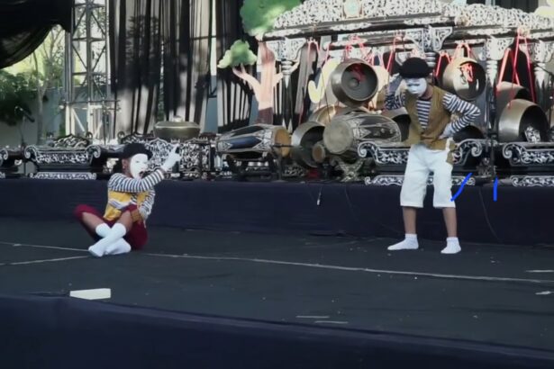 Mime with white makeup, striped shirt and yellow vest sits on stage, gesturing to a standing performer in white shorts beside ornate drums on a decorated stage.