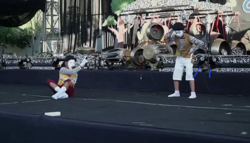 Mime with white makeup, striped shirt and yellow vest sits on stage, gesturing to a standing performer in white shorts beside ornate drums on a decorated stage.