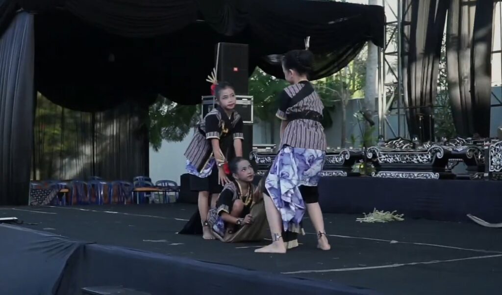 Three women in traditional attire perform a dance on an outdoor stage, one kneeling while two stand nearby, holding poses amid black stage drapery and decorative props on stage.
