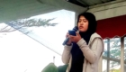 Woman in a black hijab and beige jacket speaks into a handheld microphone, looking at her phone during a talk outdoors.