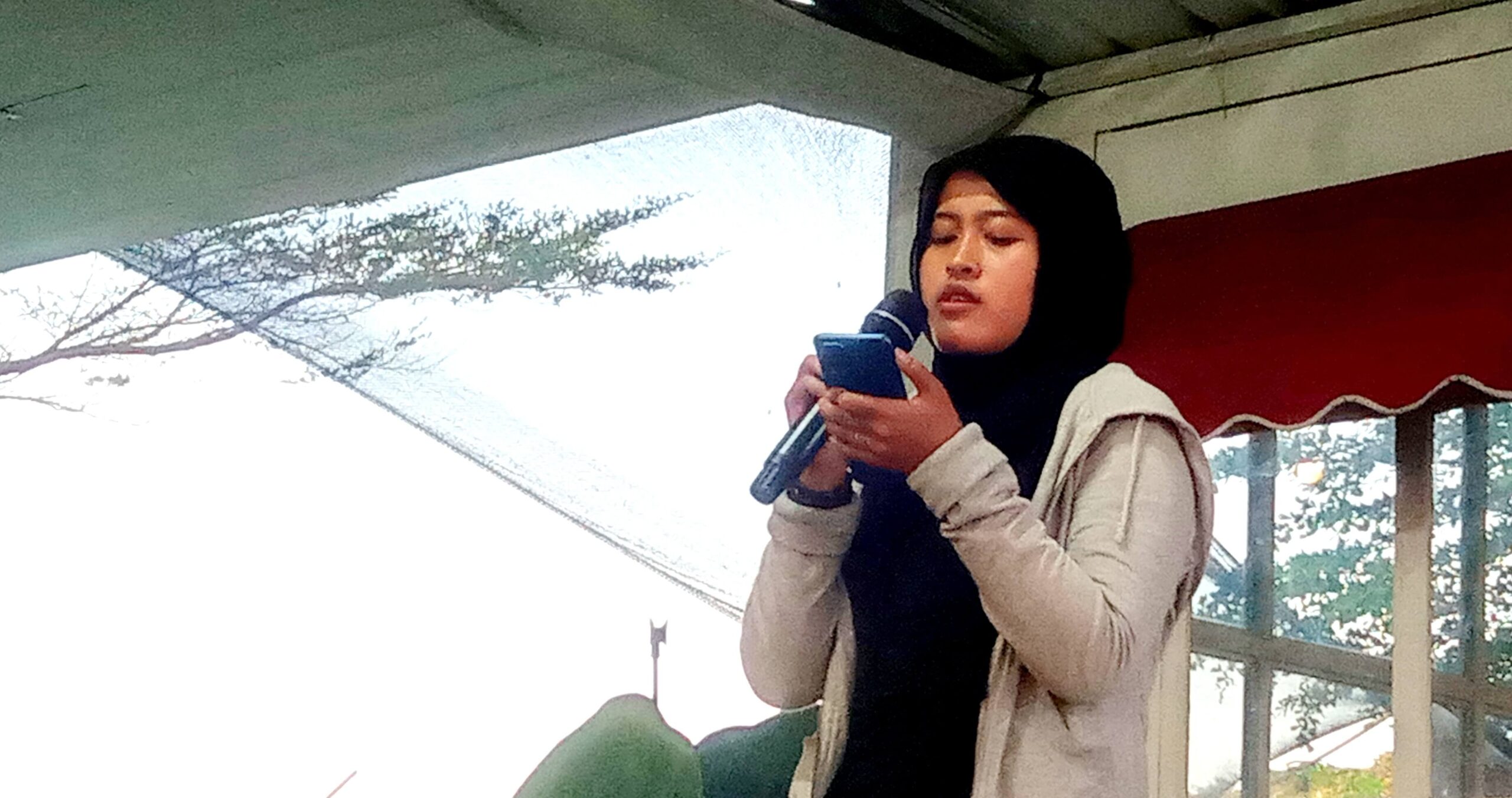 Woman in a black hijab and beige jacket speaks into a handheld microphone, looking at her phone during a talk outdoors.