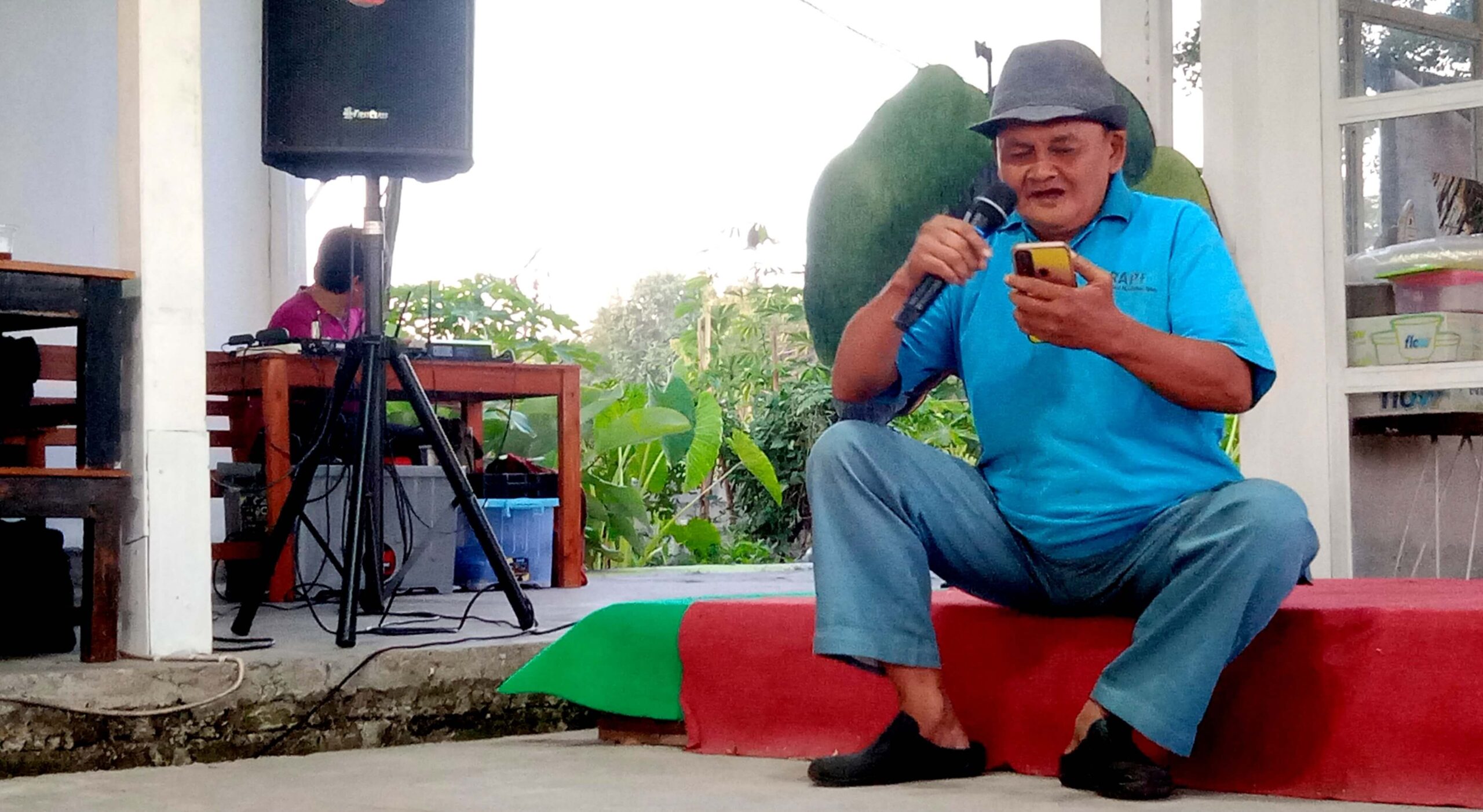 Older man in a blue shirt and hat kneels on a red carpet, speaking into a handheld microphone while looking at a yellow smartphone.
