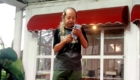 Older man with long gray hair tied back, wearing a black T-shirt, holding a microphone and a smartphone, standing in front of glass doors.