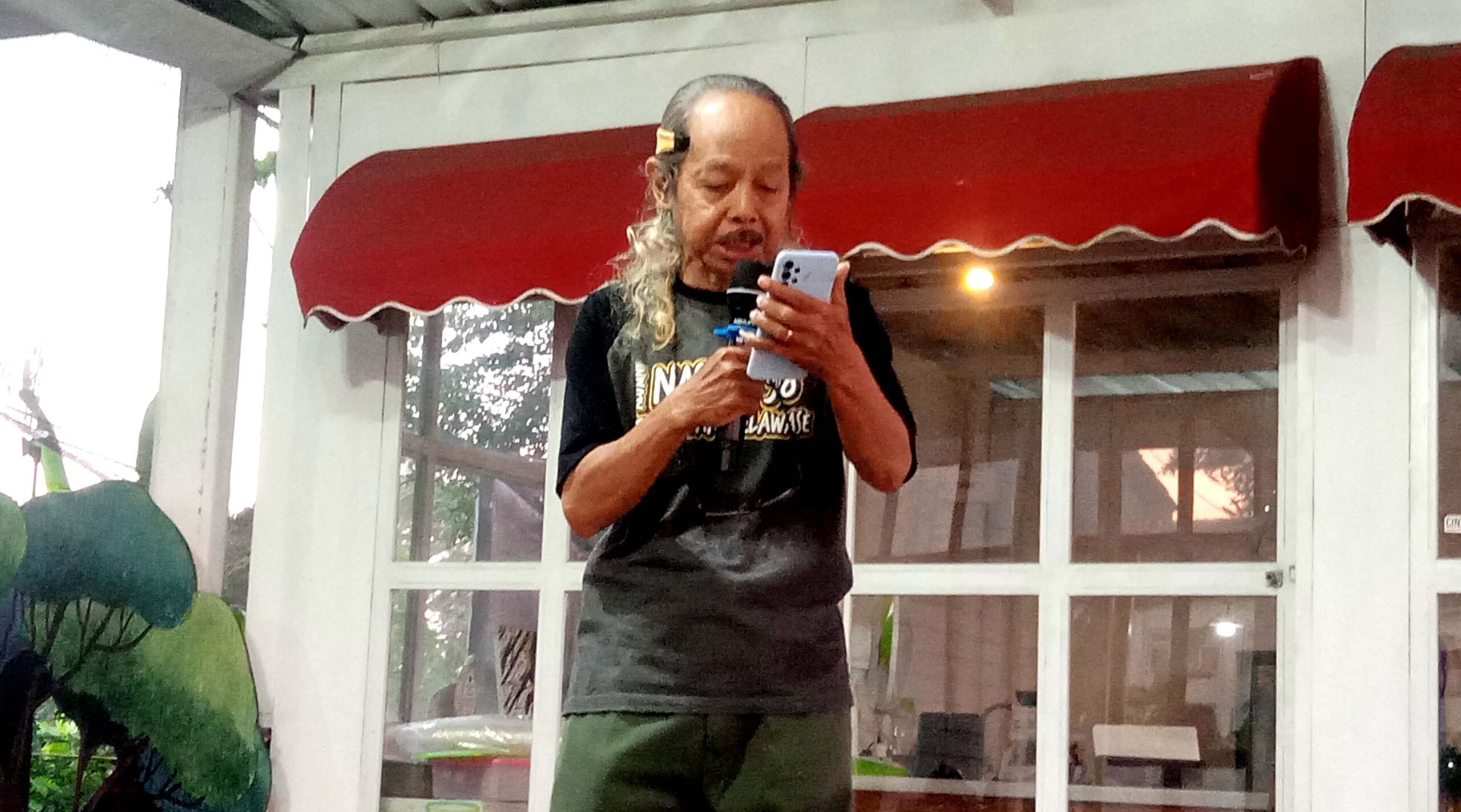Older man with long gray hair tied back, wearing a black T-shirt, holding a microphone and a smartphone, standing in front of glass doors.