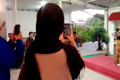 Speaker wearing a hijab and patterned dress speaks into a microphone on a small stage, holding her notes as an audience watches and records with phones.