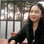 Smiling woman with dark hair in a black top sits at a wooden table indoors, sunglasses hanging from her shirt, window and trees behind her.