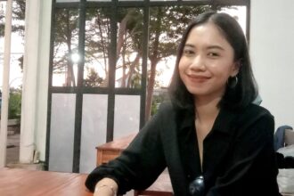 Smiling woman with dark hair in a black top sits at a wooden table indoors, sunglasses hanging from her shirt, window and trees behind her.
