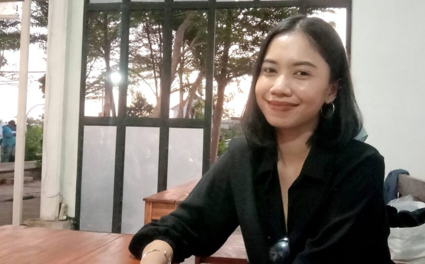Smiling woman with dark hair in a black top sits at a wooden table indoors, sunglasses hanging from her shirt, window and trees behind her.