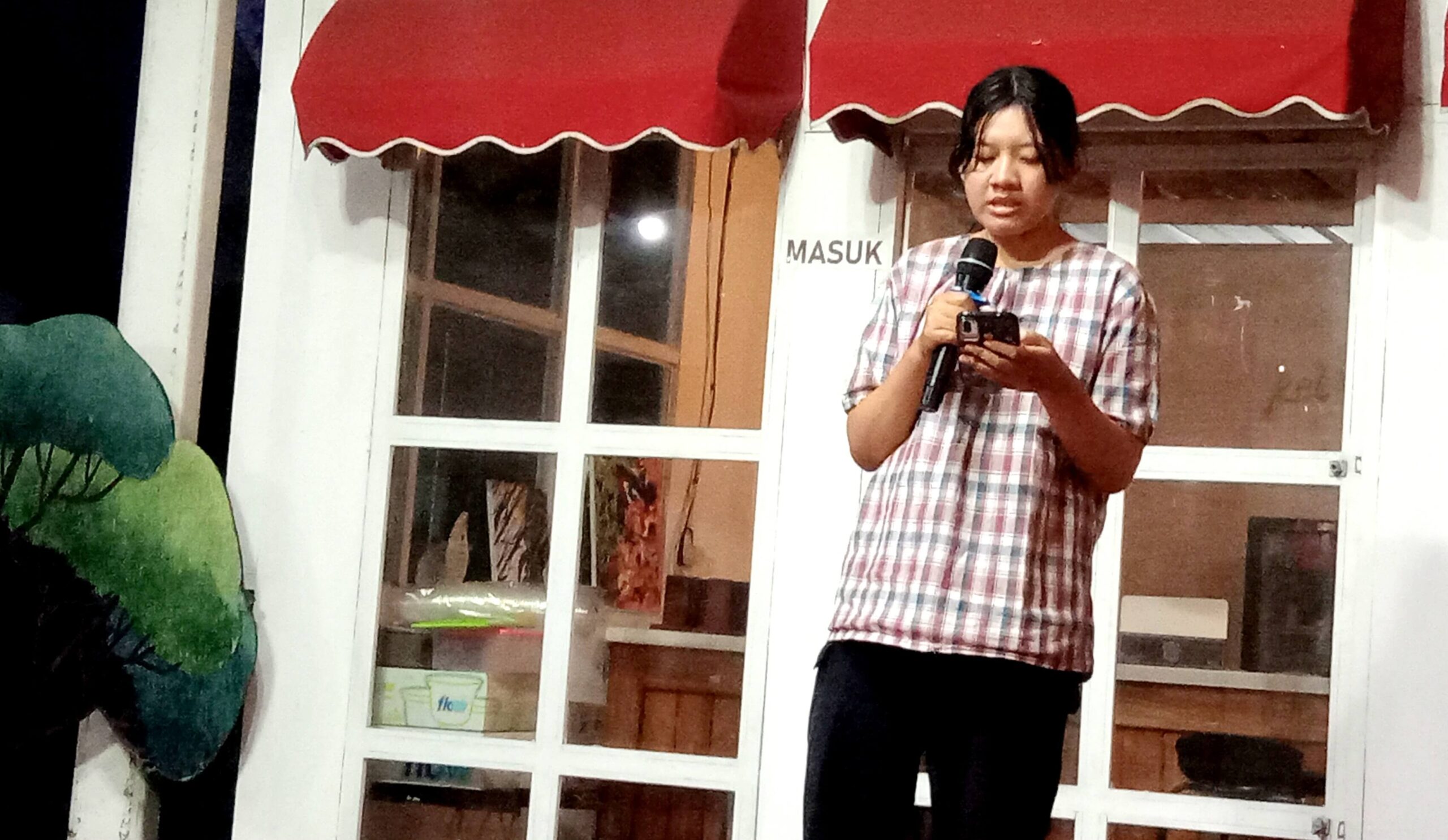 Person holding a microphone and a phone, speaking outdoors in front of glass doors, wearing a plaid shirt.