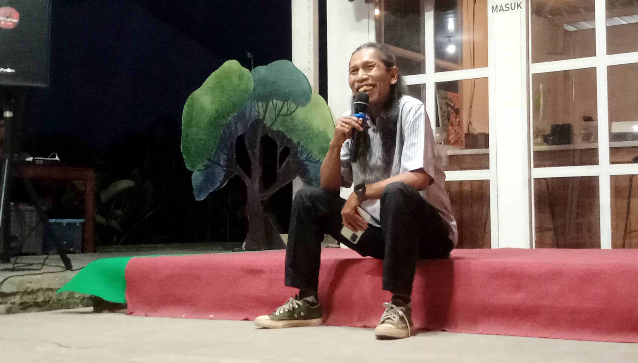 Man kneeling on a pink stage, speaking into a handheld microphone with a smile.