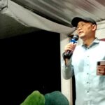 Man in a light blue shirt and dark cap speaks into a microphone on a stage, holding a smartphone in his other hand.