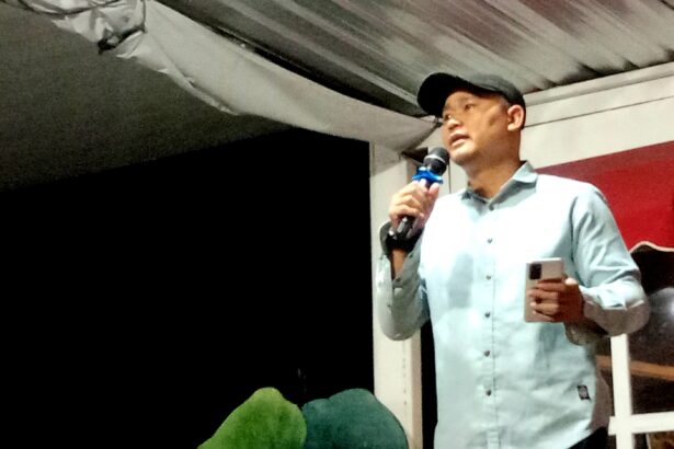 Man in a light blue shirt and dark cap speaks into a microphone on a stage, holding a smartphone in his other hand.