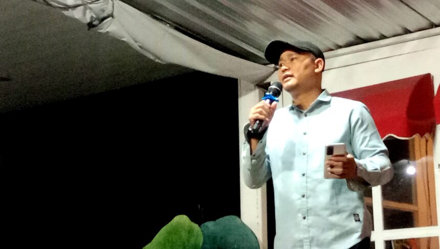Man in a light blue shirt and dark cap speaks into a microphone on a stage, holding a smartphone in his other hand.