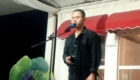 Man singing into a microphone on a small outdoor stage at night, wearing a black shirt.