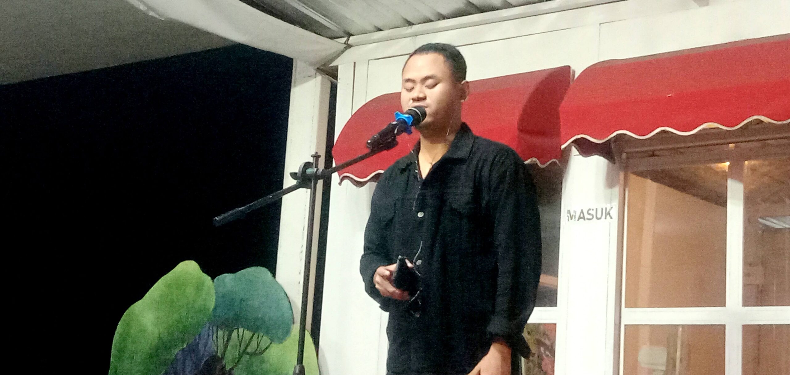 Man singing into a microphone on a small outdoor stage at night, wearing a black shirt.