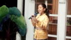 Girl wearing a gold traditional outfit speaks into a handheld microphone, holding a card, near a door with a MASUK sign in the background.