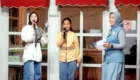 Three young women in hijabs sing on a stage, each holding a microphone, smiling at the audience.