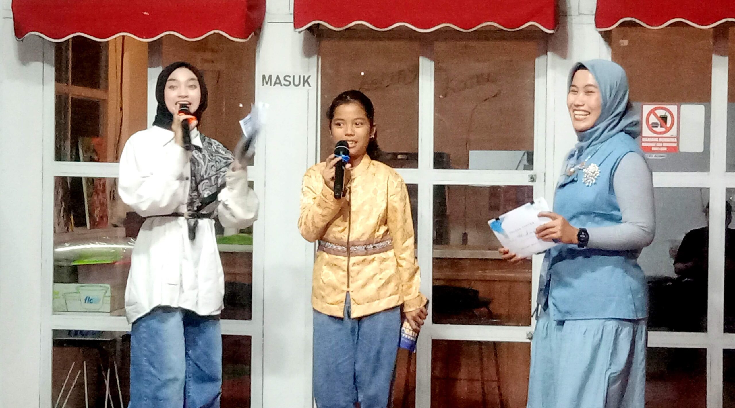 Three young women in hijabs sing on a stage, each holding a microphone, smiling at the audience.