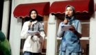 Two women in hijabs standing with microphones and notes on a stage with red canopies, presenting or hosting an event.
