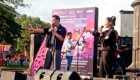 Two performers on a stage singing into microphones, with a large purple event banner behind them reading MotoGP Indonesia 2026 and a smiling crowd in the background.