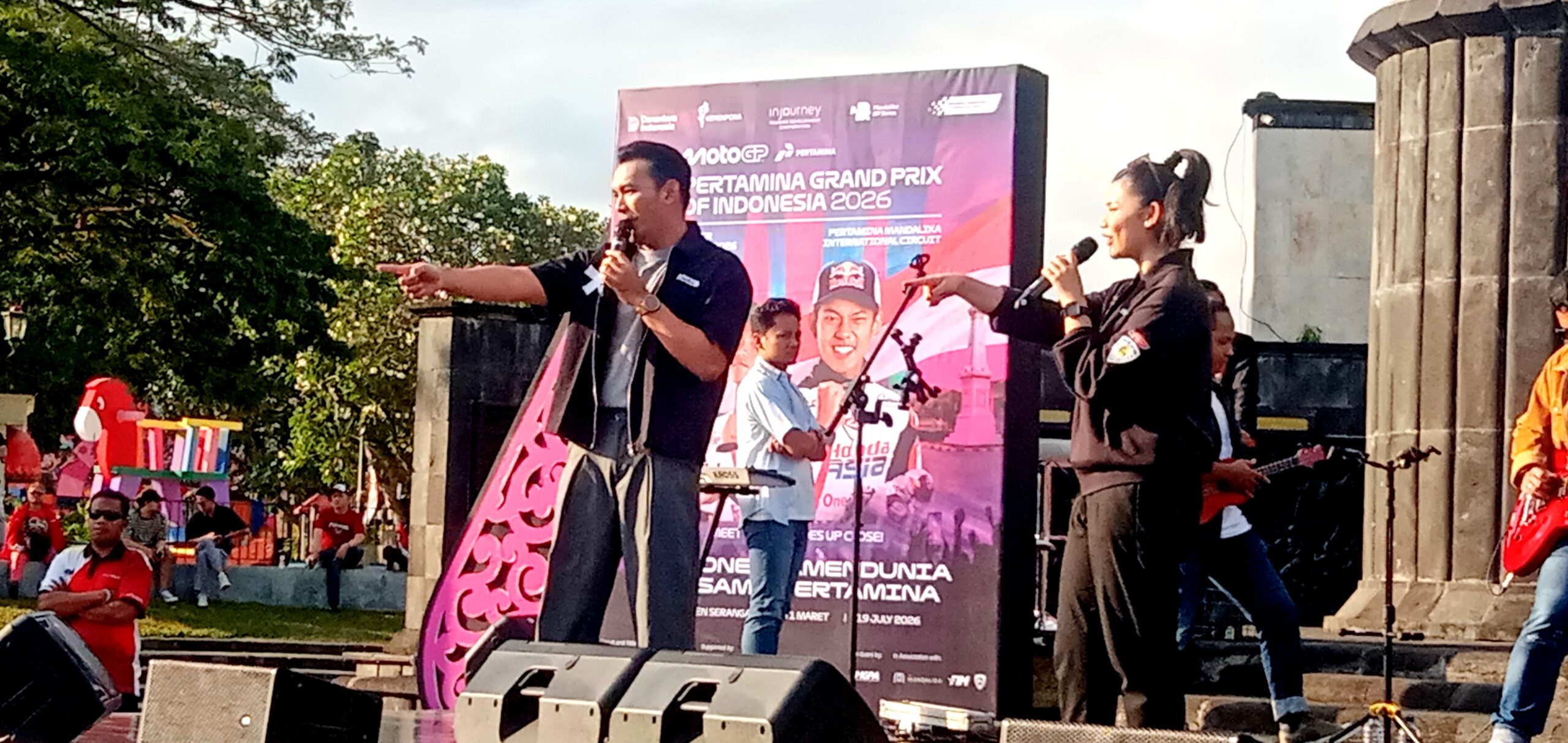 Two performers on a stage singing into microphones, with a large purple event banner behind them reading MotoGP Indonesia 2026 and a smiling crowd in the background.