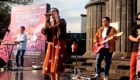 Live band performing on an outdoor stage with a female singer center, guitarist to the right, and bassist on the far right, in front of a pink banner poster.