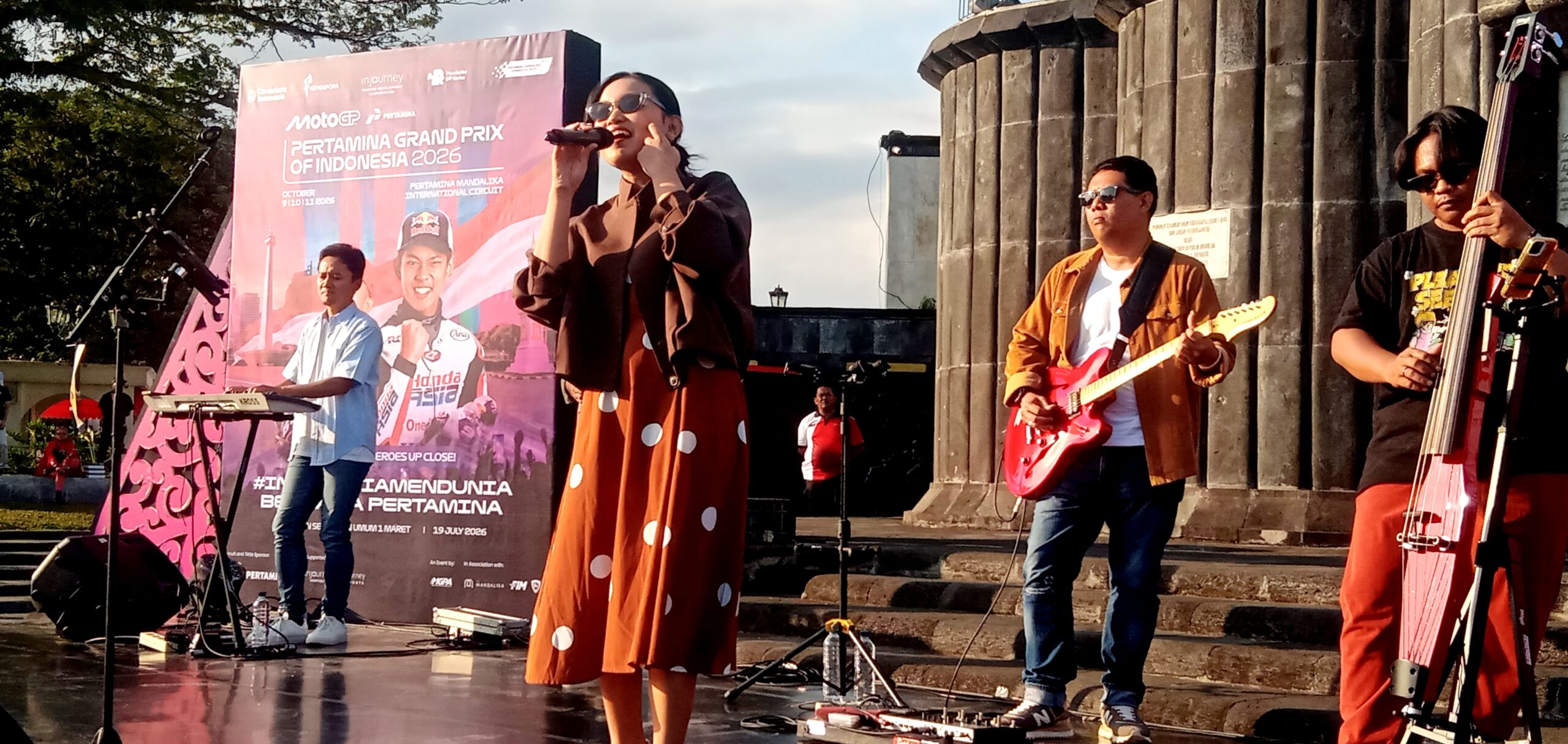 Live band performing on an outdoor stage with a female singer center, guitarist to the right, and bassist on the far right, in front of a pink banner poster.