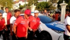 Group of security personnel in red and white uniforms near a police car at a fenced public event, with onlookers in the background.