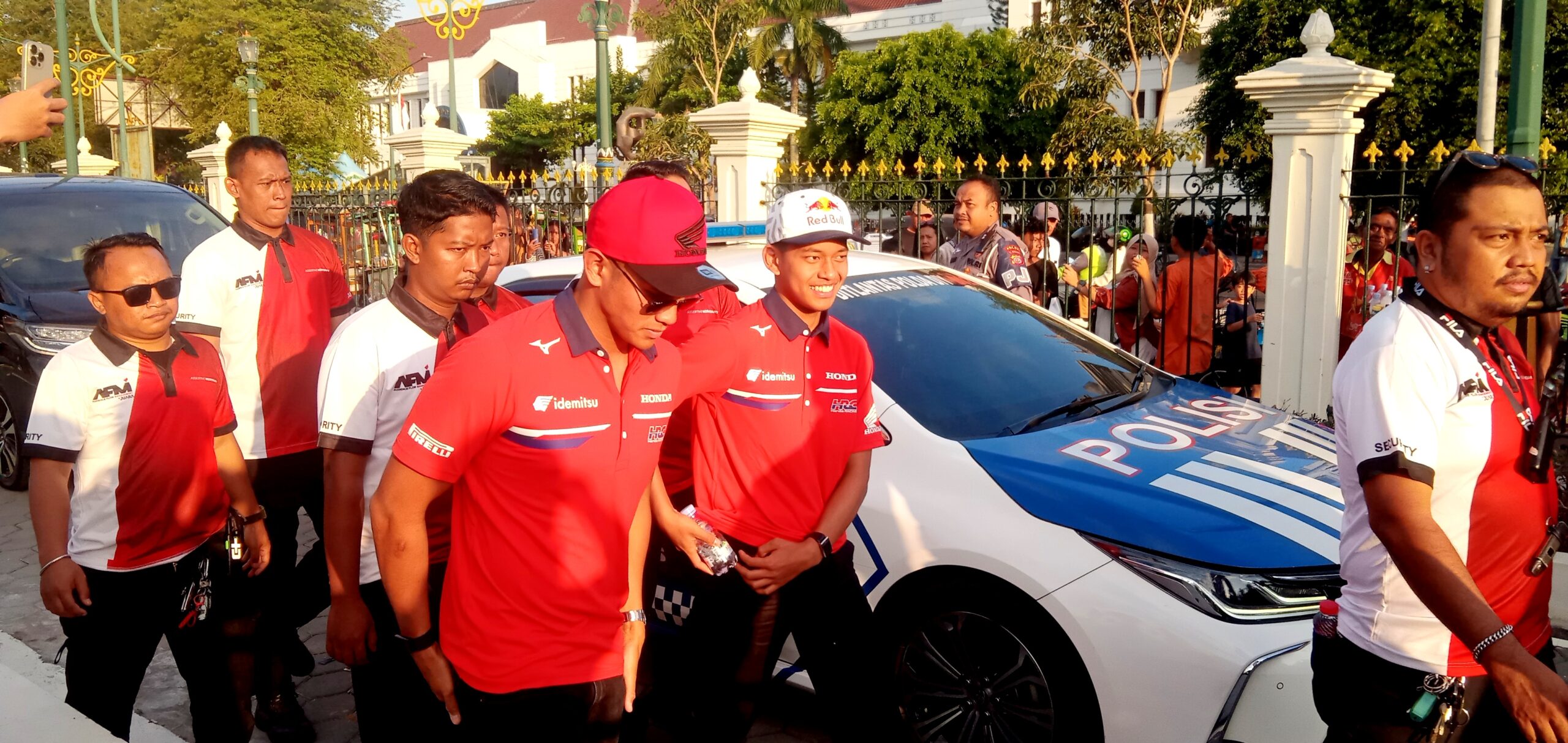 Group of security personnel in red and white uniforms near a police car at a fenced public event, with onlookers in the background.