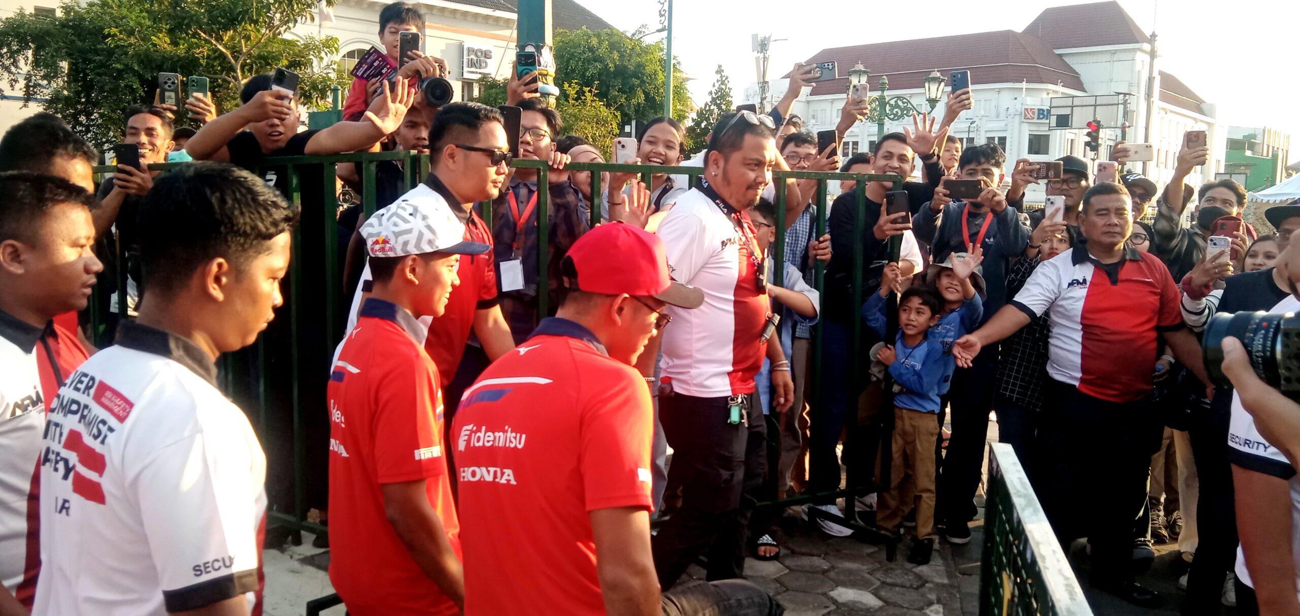 Crowd of spectators behind a green fence taking photos and videos of athletes in red and white jerseys at an outdoor event.