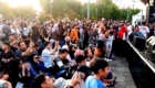 Crowd gathered at an outdoor event, many people holding up phones to take photos, stage on the right.