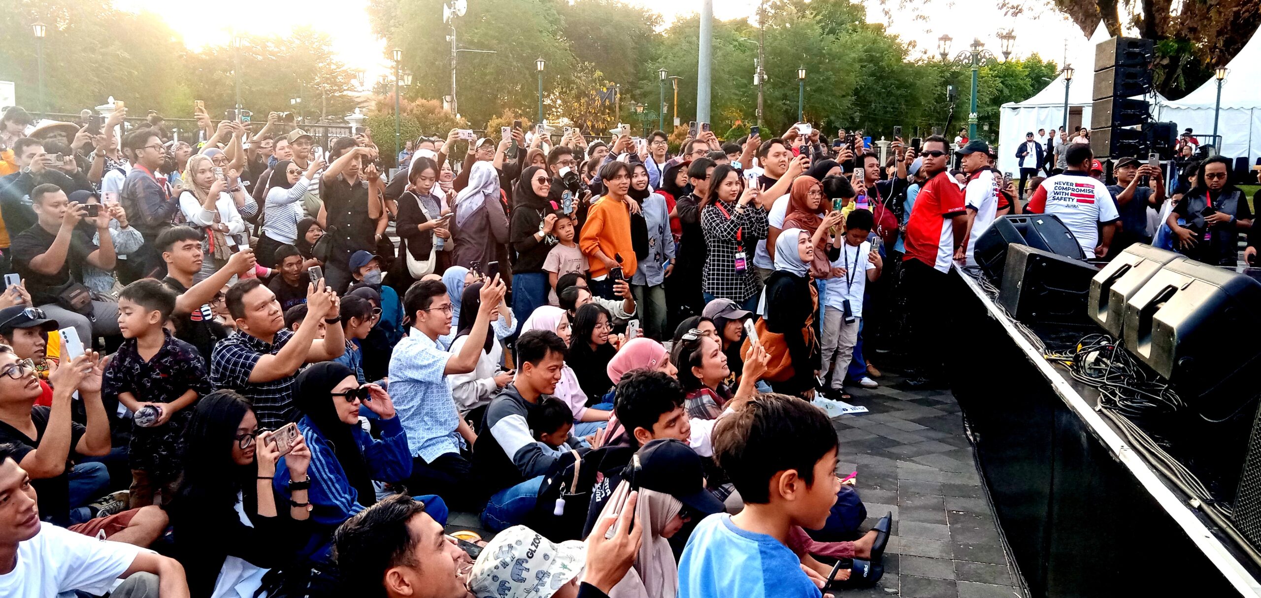 Crowd gathered at an outdoor event, many people holding up phones to take photos, stage on the right.