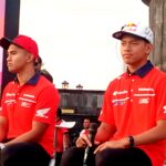 Two men in red Honda shirts sit on a stage during a motorsport event, with a colorful banner behind them.
