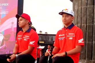 Two men in red Honda shirts sit on a stage during a motorsport event, with a colorful banner behind them.