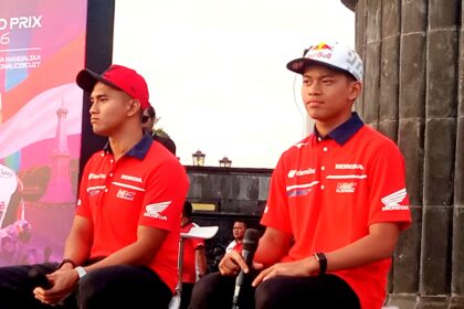 Two men in red Honda shirts sit on a stage during a motorsport event, with a colorful banner behind them.