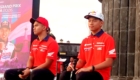 Two men in red Honda shirts sit on a stage during a motorsport event, with a colorful banner behind them.