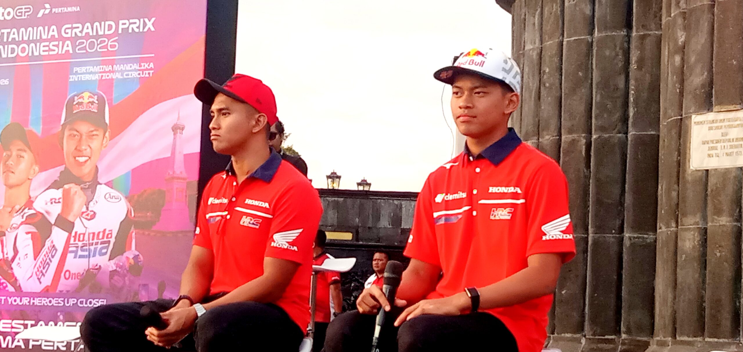Two men in red Honda shirts sit on a stage during a motorsport event, with a colorful banner behind them.