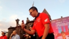 Man in a red Honda shirt and sunglasses takes a selfie with his phone at a MotoGP Indonesia 2026 event, with statues and a decorative backdrop behind him.