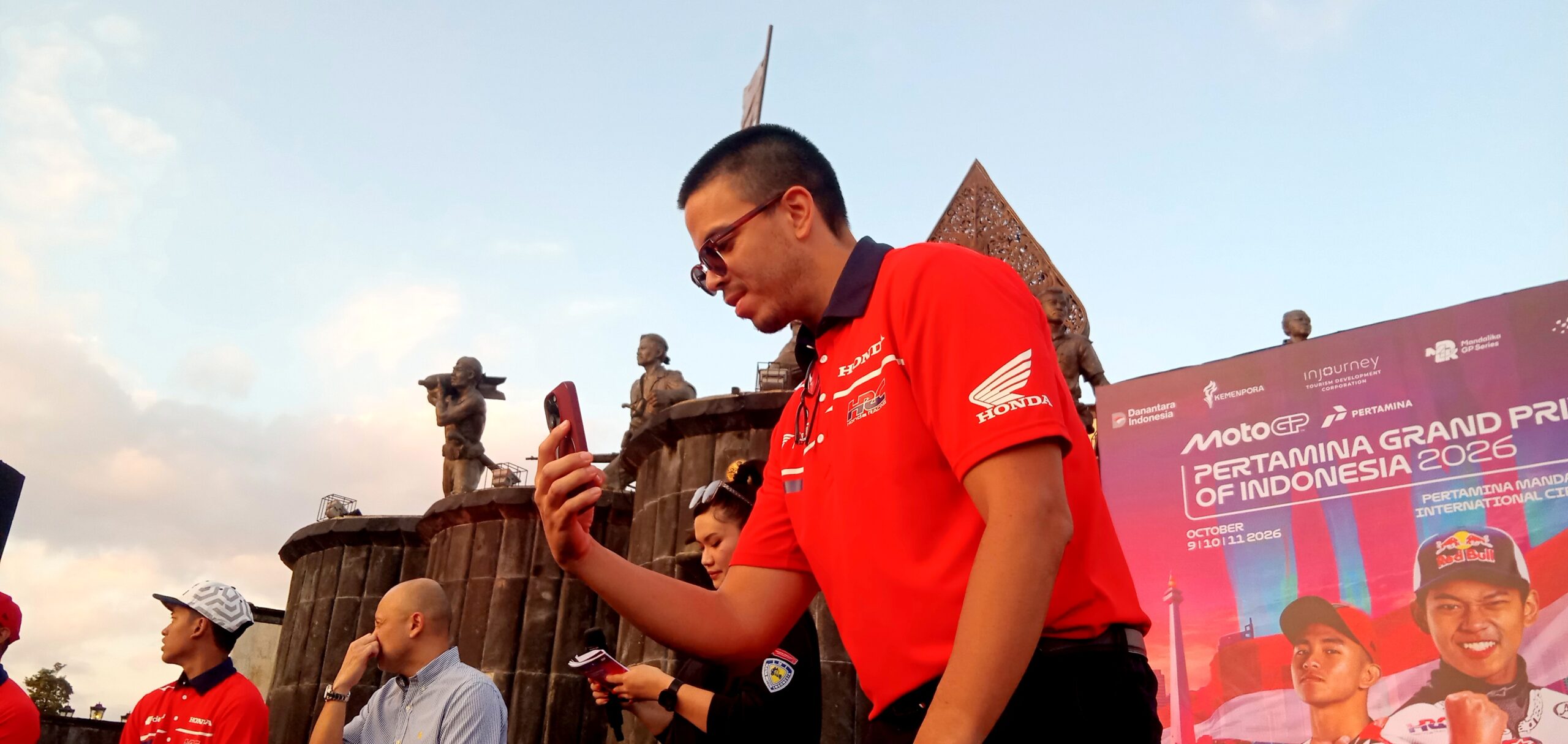 Man in a red Honda shirt and sunglasses takes a selfie with his phone at a MotoGP Indonesia 2026 event, with statues and a decorative backdrop behind him.