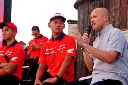 Man in a striped shirt speaks into a handheld microphone on stage, with three men in red Honda shirts nearby against a dark wall backdrop.