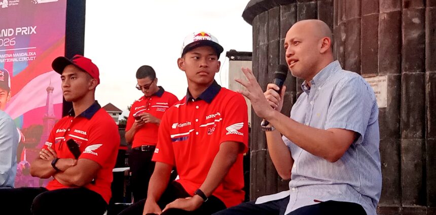 Man in a striped shirt speaks into a handheld microphone on stage, with three men in red Honda shirts nearby against a dark wall backdrop.