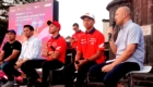 Group of men seated on a stage during a motorsport event, with a large pink banner in the background and a speaker holding a microphone on the right.
