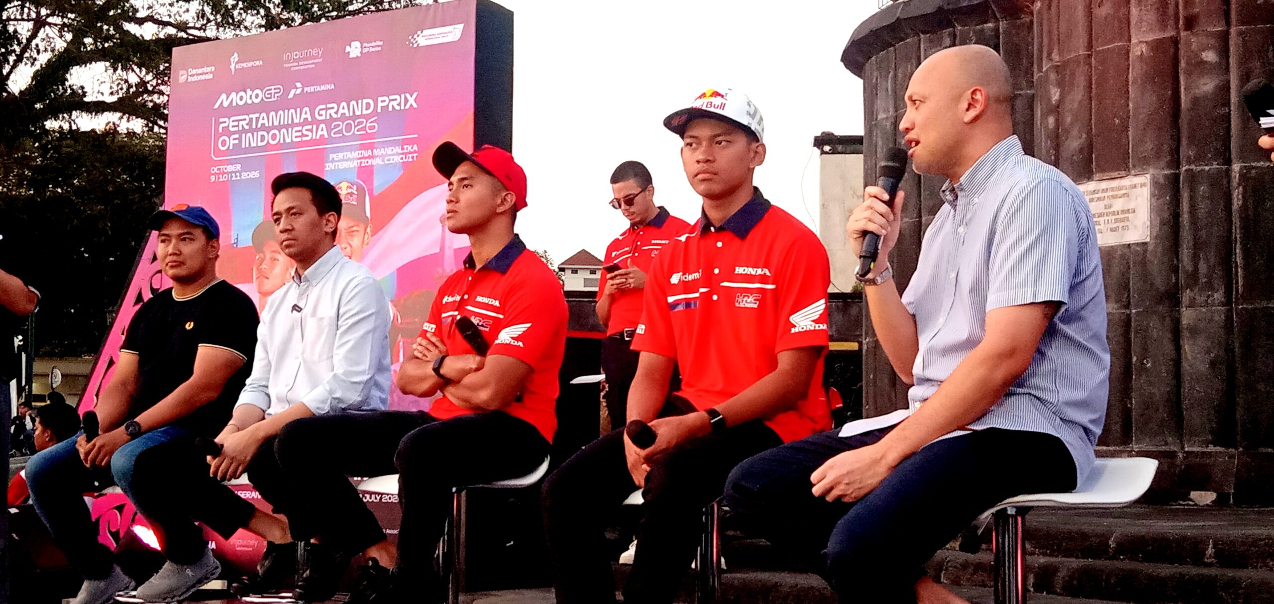 Group of men seated on a stage during a motorsport event, with a large pink banner in the background and a speaker holding a microphone on the right.