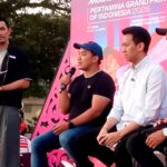 Four men sit on a stage at an outdoor event; one man in a black shirt holds a microphone while others listen, with a bright pink promotional backdrop behind them.
