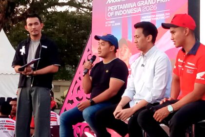 Four men sit on a stage at an outdoor event; one man in a black shirt holds a microphone while others listen, with a bright pink promotional backdrop behind them.